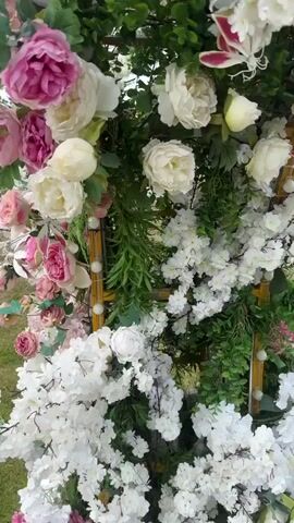 This is a close-up, vertical, slow-motion video of an outdoor floral arrangement, likely a wedding or event decoration. The arrangement features an abundance of artificial flowers and greenery. Dominant colors include various shades of pink, white, and deep green foliage. Pink peonies and hydrangeas are prominent, alongside clusters of small white blossoms, possibly cherry blossoms. The greenery consists of dense, rounded leaves, creating a lush backdrop. Interspersed throughout the arrangement are thin, gold-colored decorative elements, possibly bamboo or reeds, with small, clear decorative orbs attached. The video pans slowly upwards and slightly to the right, revealing more of the intricate floral display. The background is blurred, but suggests an outdoor setting with hints of sky and possibly grass at the very bottom of the first frame. There are no people visible in the video, and the focus remains entirely on the static floral decoration.