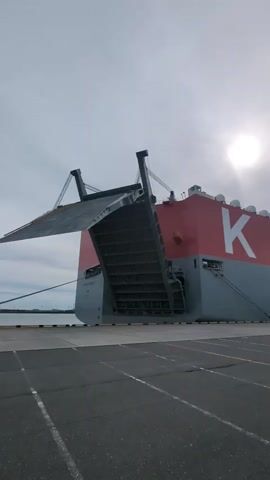 This video shows the stern ramp of a large red and gray car carrier ship. The ramp is partially lowered, extending out over the water and connecting to a dock. The ship has a large white "K" logo on its side. The sky is overcast with the sun visible through the clouds. The water is calm.