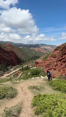The video shows a scenic mountainous landscape under a bright blue sky with scattered white clouds. The prominent feature is a large, layered red rock formation on the right side of the frame, contrasting with the green vegetation covering parts of the mountains and valleys.

In the mid-ground, three individuals are standing on a dirt path overlooking the valley. They appear to be hikers or tourists, observing the panoramic view. One person, wearing a dark jacket and backpack, is holding up a device, possibly taking a photograph or video of the scenery. The other two are standing beside them, one in a red top and the other in dark clothing.

The valley below features more green hills and scattered trees, with a winding road visible along its floor. In the distance, layers of mountains fade into the horizon. The overall impression is one of natural beauty and vastness, typical of the Jeti-Ögüz region in Kyrgyzstan, known for its distinct red sandstone formations. The time of day appears to be daytime, with ample sunlight illuminating the landscape.