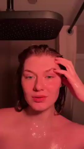 A light-skinned woman with reddish-brown hair is taking a shower. She has water in her hair and on her face. The shower head is a large, rectangular, dark color. The light appears to be red, which gives the woman’s skin and hair a red tinge. A shower curtain is slightly visible. The woman is looking around and speaking to the camera. She touches her forehead and licks her finger. A song is playing in the background.