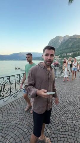 A man in a brown shirt and black shorts is standing on a cobblestone path next to a body of water. He is holding a phone in his left hand and gesturing with his right hand. In the background, there are mountains, trees, and several other people walking along the path. The sky is a clear blue. The man appears to be speaking to the camera and is possibly vlogging or recording a message.