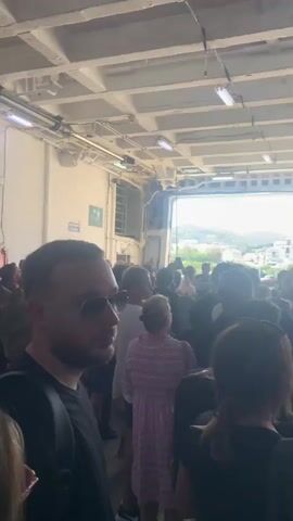A group of people are gathered on a ferry. The ferry appears to be in a port, with a town visible in the background. The people are standing and some are looking out towards the town. The video is shaky and some of the faces are blurred.