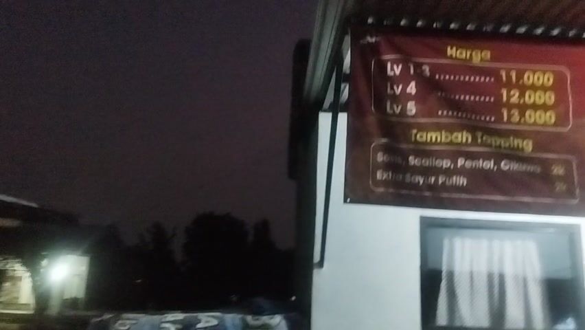 The media file captures a close-up view of a red banner displaying a menu, likely for a food stall or restaurant. The banner is mounted outdoors, possibly under a corrugated metal roof, suggesting an open-air setting. It is nighttime, indicated by the dark sky and the artificial lighting illuminating the banner and a portion of the building's wall. The weather appears calm, with no visible signs of rain or strong winds.

The banner is primarily red with yellow and white text. At the top, the word "Harga" (Price) is prominently displayed. Below this, there are listings for different levels of spiciness, "Lv 1-3," "Lv 4," and "Lv 5," with corresponding prices of 11,000, 12,000, and 13,000 respectively. Underneath the price list, the heading "Tambah Topping" (Add Topping) appears. Under this, two options are listed: "Sosis, Scallop, Pentol, Cikuwa" (Sausage, Scallop, Meatball, Cikuwa) and "Extra Sayur Putih" (Extra White Vegetable), both priced at 2k (likely 2,000). To the right of the text, there are three images of food: one appears to be noodles with toppings, another a dish labeled "Mie Hitam Pedas, Asin, Gurih" (Black Noodles Spicy, Salty, Savory), and a third showing dumplings or fried wontons labeled "Pangsit Goreng Ayam" (Fried Chicken Wontons). A logo with a cartoon character and the text "Spicy Noodles88" is also visible.

The building beneath the banner has a white wall and a dark-framed window with white curtains. A door is also visible to the right, with a small "TUTUP" (CLOSED) sign hanging on it, suggesting the establishment might be closed or about to close. The overall mood is utilitarian, focusing on the menu and pricing. The location is identified as Prajuritkulon, Indonesia.