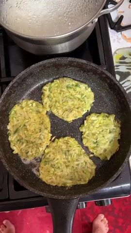 The video shows a close-up overhead view of four zucchini fritters being fried in a black non-stick skillet on a stovetop. The fritters are golden brown and appear to be made with shredded zucchini and herbs. There is some oil in the pan. A pot with a lid is visible in the background, also on the stovetop. The stovetop is black and has visible burners. A red rug is partially visible at the bottom of the frame, along with two bare feet. The lighting suggests an indoor setting, likely a kitchen.