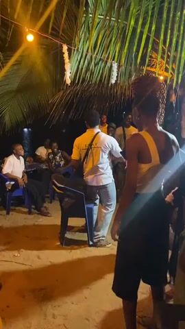 A group of young people are gathered outdoors at night. They are dancing to music. There is a chair in the middle, around which several people dance, including one man who briefly stands on the chair. Some of the dancers are wearing white shirts with designs. A small group of people are seated and watching the dancers. There are palm fronds overhead and string lights are visible. The scene takes place on a sandy area, possibly a beach.