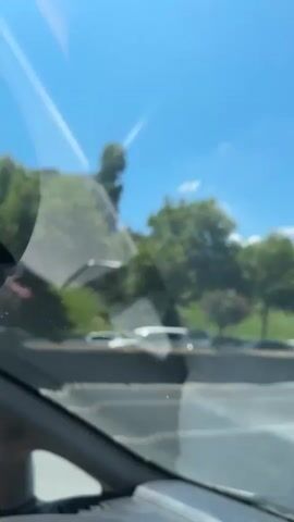 The video is shot from inside a vehicle, looking out the windshield. It shows a highway with traffic in clear daylight under a bright blue sky with a few white clouds. Several cars, including a yellow taxi, a white van, and a blue car, are visible on the road. Trees and some buildings are present on the side of the highway. The sun is shining brightly, creating lens flares and reflections on the windshield.