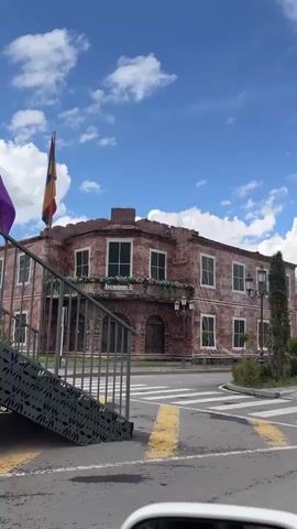 The video shows a street view with buildings and a blue sky with white clouds. On the left, there is a red brick building with white-framed windows and a balcony decorated with plants. A flag is visible on a pole to the left of the building. In front of the buildings, there is a crosswalk with yellow and white markings. The camera pans to the right, revealing more buildings with colorful murals and trees. One building has a mural of children and plants. There are streetlights and parked cars visible. The overall scene depicts a street in a town or city on a cloudy day.