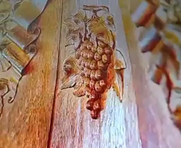 The video shows a close-up of a wooden carving of a bunch of grapes. The carving is detailed, with individual grapes and leaves clearly defined. The wood appears to be aged and has a warm, brown tone. There are other carvings visible in the background, suggesting this is part of a larger decorative piece. The lighting is warm and highlights the texture of the wood and the carving. The video is brief, showing only a few seconds of the carving from slightly different angles.