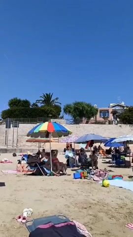 This is a video of a crowded beach on a sunny day. The camera pans across the sand, showing many people relaxing under beach umbrellas, lying on towels, and playing. Some people are in the water, wading and swimming. There are children present, some playing with toys. The background shows buildings and trees. The sky is clear and blue.
