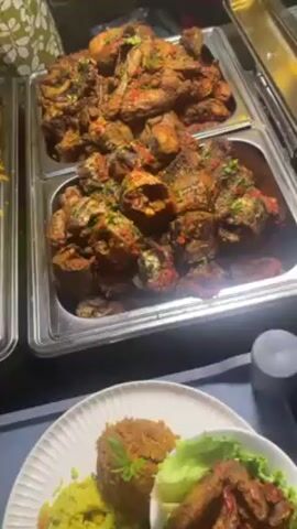 Here is a detailed description of the video.

The camera gives a close view of food laid out in a buffet arrangement. The main dishes are in large, shallow, stainless-steel trays. There is fried, reddish-brown meat covered in seasonings and red peppers. There is a tray of brown rice and a tray of bright yellow rice. There are plates of food already assembled, containing rice, pieces of meat with pepper sauce, and lettuce. There are also small bowls on the plates, filled with yellow rice. One plate also has a cooked fish, including the tail. A person in a light green patterned shirt is visible in the background.