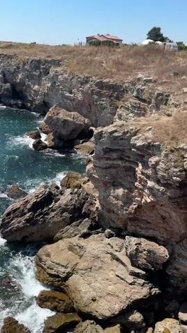 A drone captures a sweeping view of a rugged coastline with rocky cliffs and the deep blue sea. The camera pans from the rocky shore, where waves crash against boulders, to a wider expanse of the ocean. The cliffs are textured with layers of rock and sparse vegetation. In the distance, on top of the cliffs, a few vehicles and people are visible, suggesting a viewpoint or tourist area. The sky is clear and blue, indicating a sunny day.