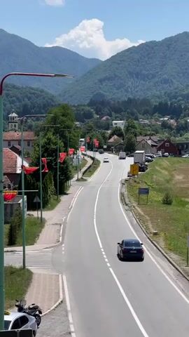 The video shows a street scene in a town nestled in a valley with lush green mountains in the background. The road is paved and curves to the right. Several cars are driving on the road. Red flags with a golden symbol are displayed on lampposts lining the left side of the road. On the left side of the road, there are houses with red roofs and a building with a white steeple, possibly a church. Further down the road, a cemetery is visible with rows of tombstones. The sky is blue with scattered white clouds. The overall impression is a peaceful rural town on a sunny day.