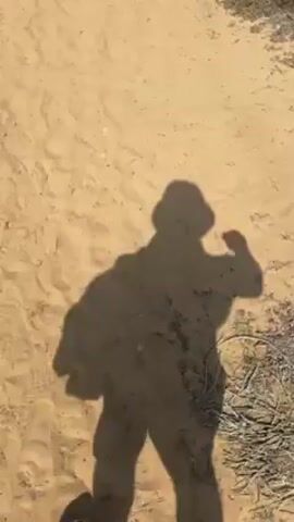 The video shows a first-person view of someone walking on a sandy path. The person is wearing sandals, and their shadow is visible on the ground, suggesting they are wearing a hat and carrying something on their back. The environment appears to be a sandy area, possibly a beach or a desert-like landscape.