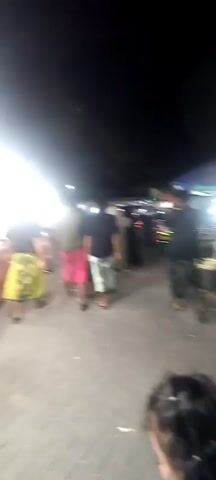 A nighttime outdoor scene shows several people walking in what appears to be a market or festival. The lighting is dim, with bright artificial lights illuminating some areas. Most people are wearing loose-fitting garments, possibly sarongs or traditional clothing. Some individuals are walking away from the camera, while others are facing different directions. In the background, there are stalls or booths, some of which are illuminated and appear to be selling goods. The ground is paved. The overall atmosphere seems casual and social.
