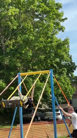 A person is performing an acrobatic routine on a swing set. The swing set is blue and yellow and is outdoors. The person is wearing a black shirt and tan pants. The person jumps from the swing and does a flip in the air before landing back on the swing. Another person is standing near the swing set and appears to be spotting the performer. There are trees in the background.