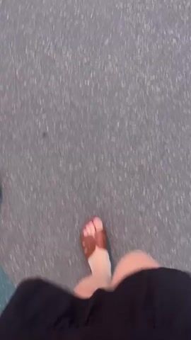 A first-person perspective video shows a person walking on a paved surface, possibly asphalt. The person is wearing brown sandals with a toe strap and dark clothing, likely shorts or a skirt. The camera is positioned to look down at the person's legs and feet as they move forward. The background is blurred due to motion, but the texture of the ground is visible, appearing to be a coarse, gray surface. The lighting suggests it is daytime, but the overall atmosphere is indistinct.