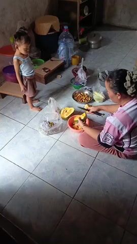 A young child stands on a small wooden stool looking down while an adult woman sits on the floor preparing food. The woman is wearing a pink and white striped shirt and pink pants. She is actively engaged in cutting or peeling a yellow-orange vegetable, likely squash, into a bowl. Surrounding them are various items, including plastic bags with food, a corn cob, a bowl of nuts or seeds, and large water jugs in the background. The environment appears to be a simple, possibly rustic, indoor space with tiled flooring and concrete walls. The lighting suggests an indoor setting, possibly during the day.