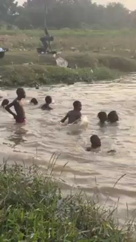 Several children are playing in a muddy river. They are dark-skinned and mostly naked, some wearing underwear. Some are submerged up to their necks, while others are splashing and running in the water. The river is surrounded by grassy banks and foliage. A lone figure is standing on the riverbank in the background, seemingly drying off or changing clothes. The lighting suggests it is either early morning or late afternoon.