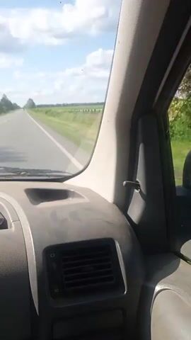 The video shows a view from inside a vehicle moving along a straight, paved road. The dashboard is visible in the foreground, with a dark gray plastic surface. A red object and a black case are on the dashboard. To the right of the dashboard, the side window of the vehicle shows the exterior. The exterior view consists of a flat, green landscape with fields and trees on either side of the road. The sky is blue with white, fluffy clouds. The video appears to be a first-person perspective of someone traveling in a car.