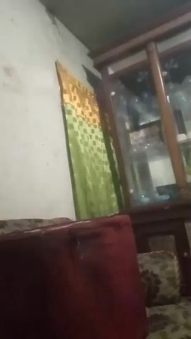 The video shows a dimly lit room with furniture and a display cabinet. A red sofa is in the foreground, partially obstructing the view of a patterned sofa behind it. A wooden display cabinet with glass doors is visible on the right side of the screen, containing several stuffed animals and what appear to be trophies or decorative items. A green and gold patterned curtain hangs on the left side of the room. The lighting is dim, creating shadows and reflections on the cabinet's glass. The camera appears to be handheld and is slightly shaky.