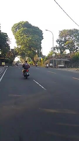 The video shows a street scene with several motorcycles and a yellow truck. The camera is mounted on a moving vehicle, likely a motorcycle, as indicated by the perspective and the movement of other vehicles. In the foreground, the asphalt road is visible with a white line marking the lane. A person on a motorcycle is seen ahead, and the vehicle moves past another motorcycle and a yellow truck in the distance. On the left side of the road, there are sidewalks, trees, and some buildings. On the right, there is a raised embankment with vegetation and a fence. The sky is bright, suggesting daylight, possibly early morning or late afternoon due to the long shadows. The overall scene depicts a normal street environment with traffic.