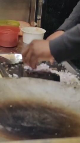 The video shows a person chopping onions on a wooden board. The person is wearing a black hoodie. In the background, there are containers of chopped onions and other ingredients. There are also some packaged food items, possibly buns, on a shelf. The scene appears to be at a food stall or street vendor, given the presence of a large cooking surface in the foreground and the general arrangement of the setup. The lighting suggests it might be evening or nighttime.