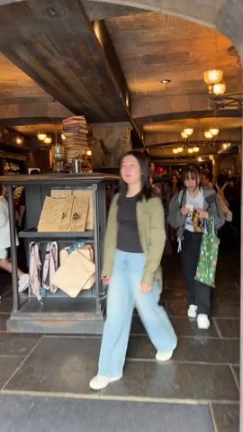 The video captures the interior of a themed retail store, likely in the Wizarding World of Harry Potter at Universal Studios Japan, given the merchandise and architectural style. Several people, primarily young adults, are seen walking through the dimly lit space. The store features stone walls, dark wooden beams and shelves, and ornate, warm-toned light fixtures.

A young woman wearing a white long-sleeved shirt over a grey vest, light blue jeans, and white sneakers, is shown walking towards the left. The camera then pans to display shelves filled with merchandise. On the left shelves are pink and white plush toys on the upper tiers, with brown pointed witch hats below. On the right shelves, there are numerous black plush toys resembling Nifflers from the Harry Potter series, along with keychains and grey bags or pouches on the bottom shelf. Another young woman, wearing a white t-shirt, dark jumper, light blue jeans, and a light-colored backpack, is seen standing in front of these shelves, looking at the products. No legible text is visible.