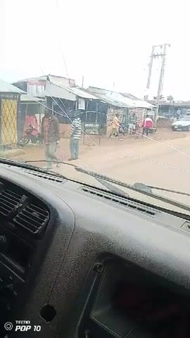The video shows a view from inside a vehicle, looking out through the windshield. Outside, there are several buildings that appear to be shops or market stalls, with corrugated metal roofs and open fronts. A dirt road passes in front of them. Several people are visible outside the buildings, and some are interacting. One person is sweeping the ground with a broom. Another person is standing near a motorcycle. There are also motorcycles and a few cars on the road. The sky is overcast, suggesting cloudy weather. A speed limit sticker is visible on the inside of the windshield at the top. The dashboard of the vehicle is also visible in the foreground. The video appears to be taken while the vehicle is either stopped or moving slowly.