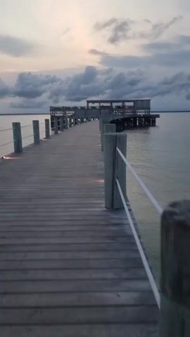 The video shows a wooden pier stretching out over a body of water toward a structure at the end. The pier has wooden posts connected by white rope railings. Small lights are built into the pier deck. The sky is overcast with clouds and the water is calm.  The pier leads to a larger platform-like structure at its end. In a brief pan to the left, a beach is visible, with some scattered people walking on it.