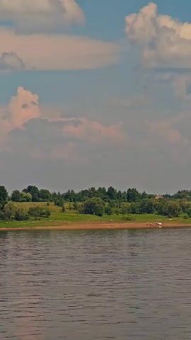 The video pans across a wide, rippling body of water, likely a river, towards a gently sloping green embankment lined with trees and bushes. On the embankment, a small cluster of rustic houses with varied rooflines is visible, suggesting a rural village or settlement. The sky above is a mix of soft blue and large, scattered clouds tinged with pinkish-orange hues, indicating either early morning or late afternoon light. The overall impression is one of a serene, pastoral landscape.