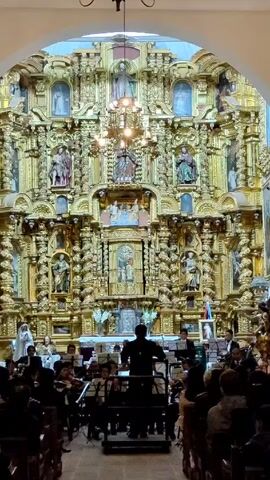 An orchestra performs in a church in front of a large golden altarpiece. The altarpiece is ornate, with numerous sculptures and images. A conductor stands in front of the orchestra, directing them. The orchestra is composed of various instruments, including violins, cellos, and woodwinds. People sit in the pews, watching the performance. The church has a high, arched ceiling. A chandelier hangs above the orchestra.