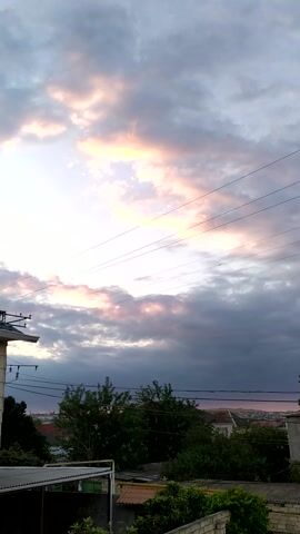 This is a vertical video clip showing a cloudy sky at sunset, as viewed from a residential area. The sun is setting, casting a warm, orange glow on some of the clouds, while others remain a dark gray. Power lines stretch across the sky, partially obscuring the view of the horizon. In the background, there are houses, trees, and some brick walls, indicating a suburban or rural setting. The overall mood is calm and peaceful, with the vibrant colors of the sunset creating a sense of beauty and serenity. The time of day is clearly sunset due to the lighting and cloud colors. There is no visible text in the video.