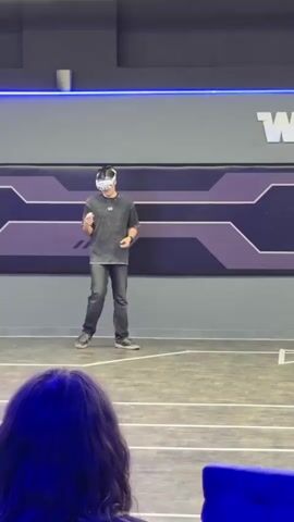 A young man wearing a VR headset stands on a marked floor, holding two controllers in his hands. He’s facing forward, looking down. In the foreground, the back of a woman’s head is visible, and she turns her head to the right to look at the man. The room has a gray wall with a purple and gray design, and the word "WART" is visible in white letters. Above the wall is a blue light strip. Overhead lights are on. The floor is marked with white lines. The woman is sitting in a blue chair. Music plays in the background.