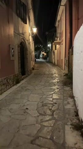 A person walks down a narrow, stone-paved alleyway at night. Buildings line both sides of the alley, illuminated by streetlights and lights from within the buildings. One building on the left has a pink facade, and a large, arched doorway. The building on the right is also pink and has balconies. The alley is dimly lit, with shadows cast by the buildings and the lights.