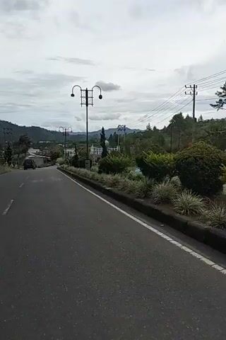 The video shows a paved road with a white line marking the edge. On the right side of the road, there is a stone border with a variety of plants, including ornamental grasses and bushes. A tall lamppost with a decorative top stands on the right side of the road. In the distance, a white building with a blue dome, possibly a mosque, is visible. A dark-colored SUV is driving away from the camera on the left side of the road. The road curves to the left and leads into a valley with hills covered in greenery. The sky is overcast with scattered clouds. The overall scene appears to be a scenic view of a road in a rural or semi-rural area.