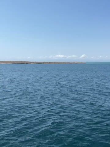 The video shows a wide expanse of blue water under a bright, pale blue sky dotted with a few fluffy white clouds. In the distance, a long, low strip of land is visible, appearing to be a coastline or island with brownish, vegetation-covered terrain. The water is rippled, indicating a slight breeze. No people or boats are visible in the foreground, and the sound of someone speaking can be heard in the background.