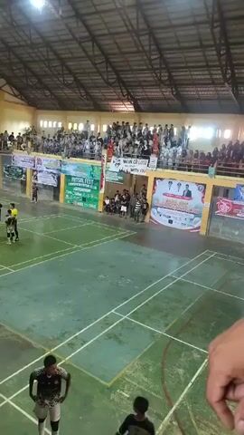 The video shows an indoor sports arena with a futsal game in progress. The court is marked with white lines for futsal and badminton. There are spectators in the stands, many of whom are young people. The lighting appears to be artificial. Several banners are visible on the walls, some with text and images. A few players are on the court, some in uniforms. One player in the foreground is looking down at the court. The camera angle is from a high vantage point, looking down at the court. The overall atmosphere appears to be energetic and crowded.
