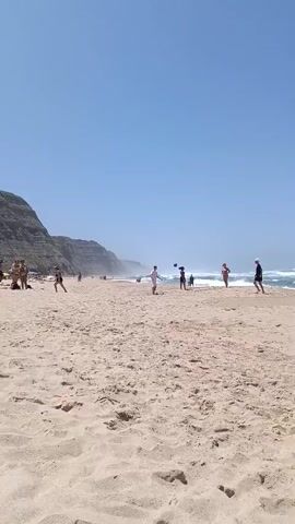 The video shows a crowded beach with light brown sand and a bright blue sky. The ocean is visible in the background with moderate waves. Several people are scattered across the beach, some walking, others standing. A group of people appears to be playing a game in the middle distance. Further down the beach, a person in a white shirt and dark shorts is playing with a ball. There are also some beach umbrellas visible on the left side of the frame. A rocky cliff face rises behind the beach on the left. The sun is shining brightly.