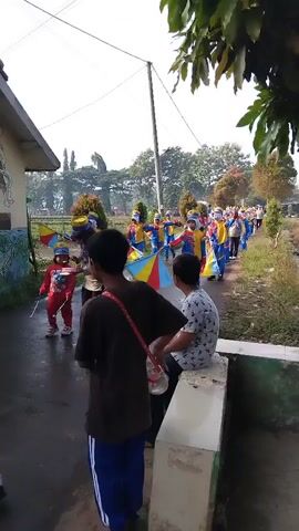 A group of children in brightly colored uniforms, some holding flags, are marching in a procession down a wet asphalt road. Two boys, one standing and one sitting, watch the parade from the side of the road. The environment appears to be rural, with trees and vegetation visible along the side of the road. The weather is overcast.