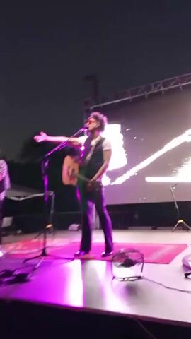 A man performs on stage with a guitar. He wears a black vest and purple trousers. He also has on a red bandana and glasses. A large screen behind him displays an image of a man with dark, curly hair and glasses. The stage is lit with pink and green lights.