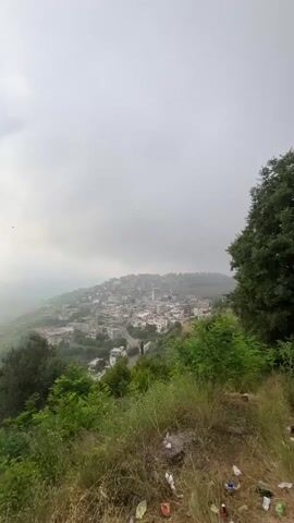 The video begins with a wide shot of a hillside town shrouded in mist. The town is densely packed with buildings of various sizes and architectural styles, appearing to be constructed primarily of concrete and stone. A prominent white minaret rises from the center of the town. The foreground is dominated by lush green vegetation, including trees and bushes, with some scattered litter visible on the ground. The camera then zooms in, providing a closer view of the town's rooftops and winding streets. The overall atmosphere is overcast and somewhat hazy.