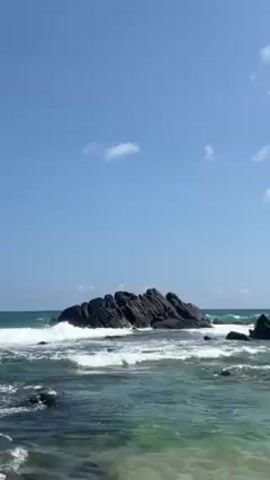 The video shows a rocky outcrop in the ocean under a clear blue sky with a few wispy clouds. Waves crash against the rocks and surrounding smaller rocks. The water is turquoise near the shore and deeper blue further out. The perspective is from the shoreline.