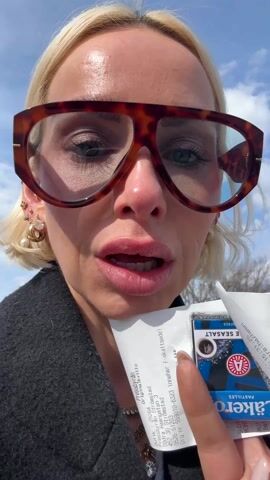 The video features a woman with short blonde hair, fair skin, and wearing oversized, tortoise-shell framed glasses. She is holding a receipt and a small package of Läkerol Salmiak Seasalts in her hand, with a blue sky visible in the background. She appears to be outside, speaking directly to the camera. The receipt shows it was purchased at Pressbyrån, a store at Södra Hamngatan 3 in Strömstad, Sweden. The woman seems to be expressing excitement about using tokens to buy the candy.