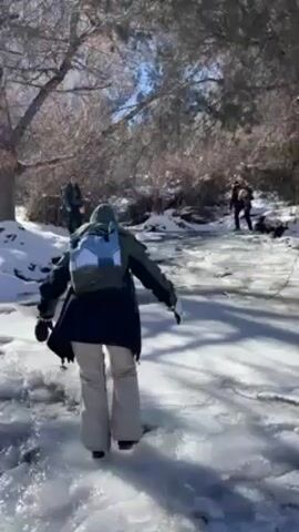 The video shows a group of people walking on a frozen, ice-covered landscape, probably a frozen stream or a water body. The person in the foreground is walking away from the camera, wearing a dark jacket, light-colored pants, and a backpack. There are other people in the distance, also walking in the same direction. The ground is covered with snow and ice. There are trees without leaves on both sides, suggesting it's winter. The scene appears to be outdoors, possibly a park or hiking trail. The sunlight reflects brightly off the ice and snow.