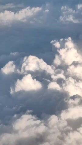 A video clip shows a view from an airplane flying above a layer of clouds. The clouds are white and fluffy with some areas of gray, suggesting varying depths and thickness. Sunlight is visible, illuminating some of the clouds and creating shadows. The sky appears blue in the upper portions of the frame. The camera appears to be moving slightly, giving a sense of aerial perspective.