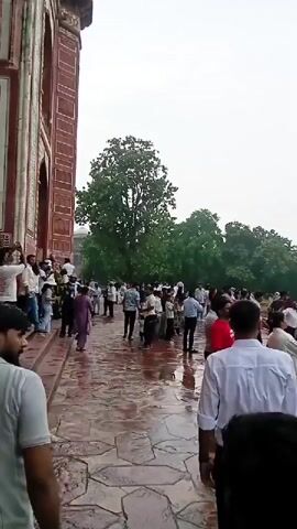The video shows a crowd of people walking through a park-like area with trees and manicured gardens. The ground is wet, indicating it may have rained or is about to rain. In the distance, a large, white domed building with minarets, which appears to be the Taj Mahal, is visible. People are dressed in casual clothing, and some are holding cameras or phones. The overall atmosphere seems to be one of tourism and sightseeing.