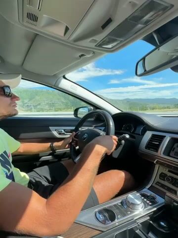 The video shows the interior of a car driving on a highway on a sunny day. The camera is positioned behind the driver, focusing on the steering wheel, dashboard, and the road ahead. The driver's hands are on the steering wheel, and their legs are visible. The dashboard features a navigation screen, air vents, and a gear shift. The road is a multi-lane highway with a concrete barrier on the left and a rocky hillside on the right. The sky is blue with scattered white clouds, and the surrounding landscape consists of hills and mountains. The car appears to be moving at a moderate speed.