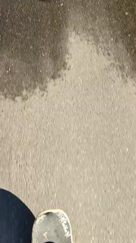 A person is riding a skateboard on an asphalt road. The view is from above, looking down at the skateboard. The road is cracked and uneven, with light reflecting off the surface. The person's foot is visible on the skateboard, wearing a black and white shoe with white laces. The camera is shaky and moves along with the skateboard.