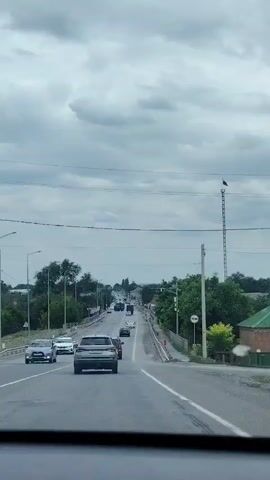 The video is a first-person view from a vehicle driving on a road. The road is paved and has multiple lanes. There are several cars driving in the same direction as the vehicle. On the right side of the road, there is a guardrail and trees. Further down the road, there is a tall pole with a flag on top and what appears to be a traffic camera. The sky is overcast with clouds.