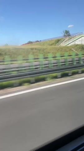 This media file captures a view from a moving vehicle, likely a car or bus, traveling along a highway. The scene is set on a sunny day with a clear blue sky, suggesting fair weather. The highway features multiple lanes marked with white lines, indicating a relatively modern road infrastructure.

On the right side of the highway, a grassy embankment rises, topped with scattered trees and shrubs. A green and metal guardrail runs along the base of the embankment, separating it from the road. In the distance, a pedestrian bridge crosses over the highway, with stairs leading up to it from the embankment. The bridge is painted green with yellow accents and has a light gray concrete support structure.

The perspective suggests the vehicle is moving at a moderate to high speed, as evidenced by the motion blur in the foliage and the background. No people are directly visible in the frames, but the presence of a highway and pedestrian bridge implies human activity and infrastructure development.

The overall mood is serene and ordinary, depicting a typical travel experience on a highway under clear skies. The location context of Choroszcz, Poland, places this scene within a specific geographical area in Eastern Europe. No visible text is present in the media file.